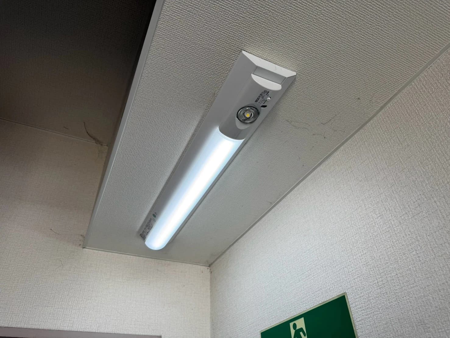 施設の廊下の天井に取り付けた非常灯内蔵の LED ベースライト。壁には避難口誘導灯