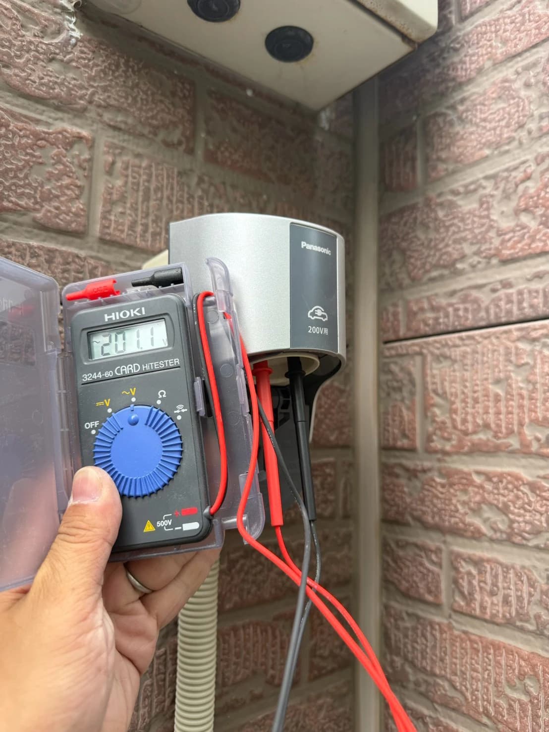 住宅の外壁に取り付けた Panasonic 製の EV 充電用 200V コンセント。テスターで 200V の電圧を確認している