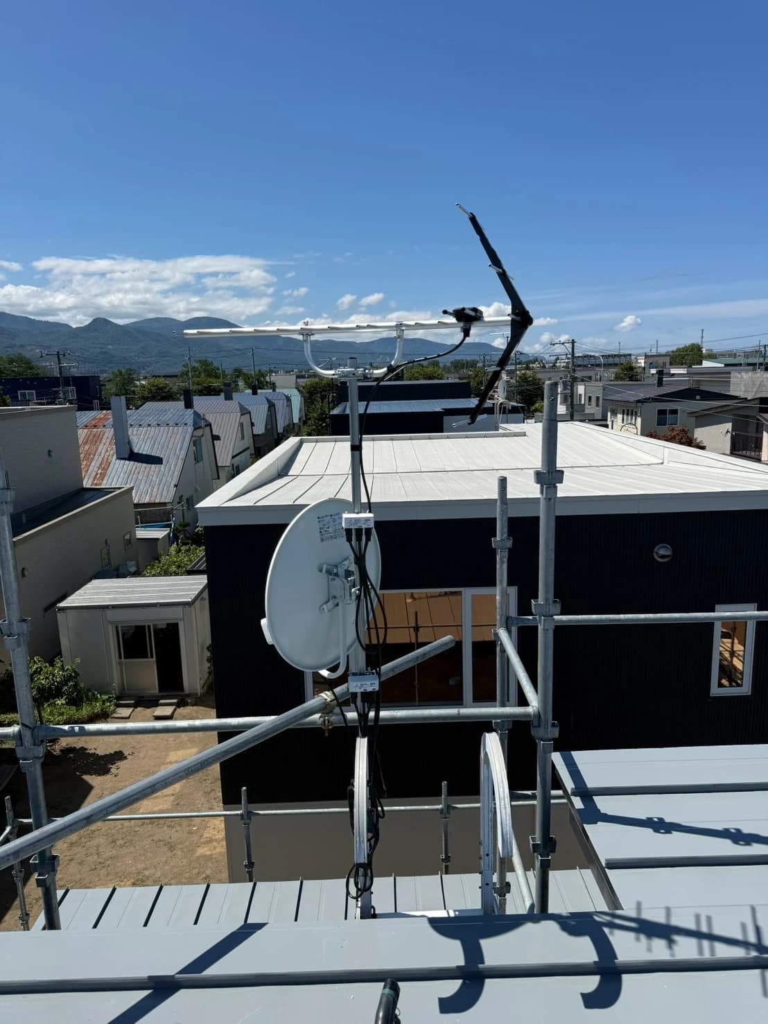 新築住宅の屋根上のマストに取り付けた UHF アンテナと BS・CS アンテナ。青空と山並みを背景に